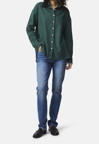 Grünes Button-up-Hemd mit entspannter Passform, kombiniert mit blauen Jeans und schwarzen Slippern. Das Hemd hat eine Frontabgesetzte und eine weiche Textur.