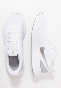 Chaussures de sport blanches avec dessus en mesh, bout rond, accents argentés et semelle texturée. Dotées de lacets et d'un détail de marque près de la languette.