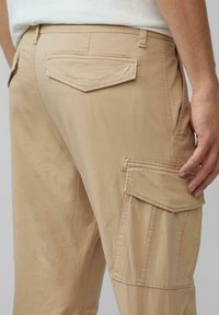 Beżowe spodnie cargo o gładkiej fakturze, z dwiema płaskimi tylnymi kieszeniami oraz dużą kieszenią cargo z boku. Szwy są widoczne wzdłuż krawędzi.