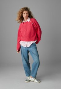 Lyserød strikket trøje med ribbet kant og sidelukninger, lagdelt over en hvid og rødstribet skjorte, parret med lyseblå jeans og sølvfarvede sneakers.