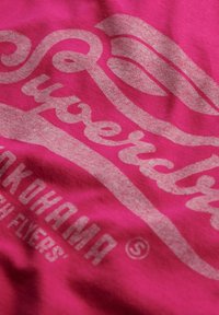 T-shirt fuchsia avec un grand graphisme gris portant le texte "Superdry" et "Yokohama" dans des polices stylisées, mettant en avant une texture douce.