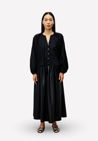 Frau mit langen schwarzen Haaren, die eine schwarze Bluse mit Knöpfen, einen schwarzen glänzenden Knielangen Rock und metallisch goldene Ballerinas trägt, aufrecht stehend.