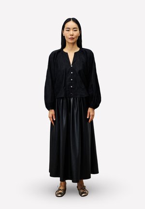 Femme avec de longs cheveux noirs portant une blouse noire boutonnée, une jupe noire brillante jusqu'aux chevilles et des ballerines dorées métalliques, se tenant droite.