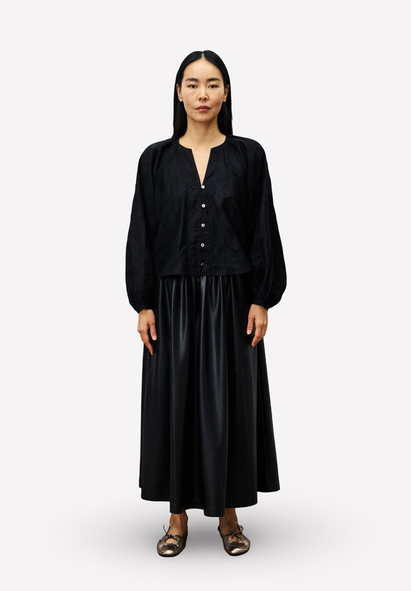 Frau mit langen schwarzen Haaren, die eine schwarze Bluse mit Knöpfen, einen schwarzen glänzenden Knielangen Rock und metallisch goldene Ballerinas trägt, aufrecht stehend.