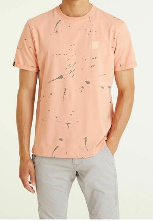 Homme portant un t-shirt rose clair avec un motif de tâches de peinture grise et un pantalon gris clair, debout avec une main dans la poche.