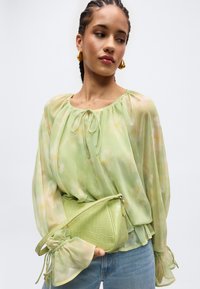 Γυναίκα που φοράει ανοιχτό πράσινο διαφανές πουκάμισο με φουσκωτά μανίκια και πράσινη υφασμάτινη τσάντα χιαστί πάνω από μπλε τζιν.