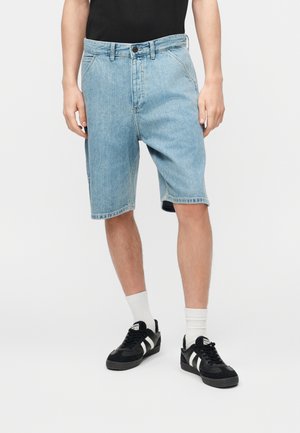 Homme portant un short en denim bleu clair avec un t-shirt noir, des chaussettes blanches et des baskets noires avec des rayures blanches sur un fond blanc.