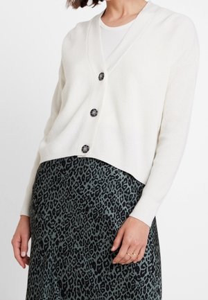 Cardigan blanc côtelé avec un col en V et cinq boutons foncés. Porté sur une jupe à imprimé léopard vert, mettant en valeur des textures et des motifs contrastés.