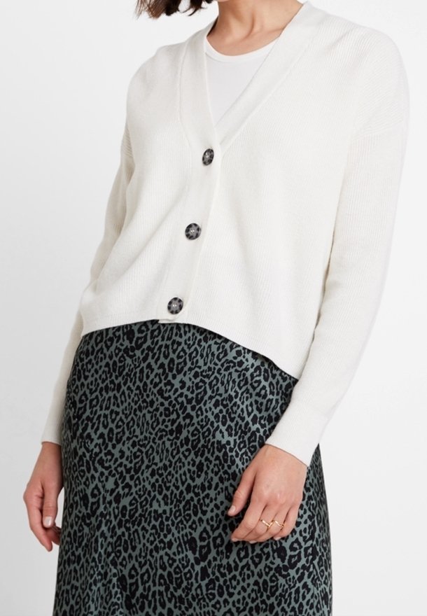 Cárdigan blanco de canalé con escote en V y cinco botones oscuros. Llevado sobre una falda verde con estampado de leopardo, mostrando texturas y patrones contrastantes.