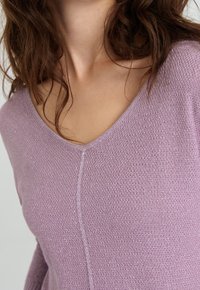 Svetle fialový svetr s výstřihem do V, strukturovaným pletením a jemným leskem, s centrálním švem a volným střihem.
