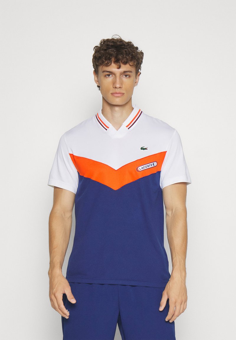 Jeune homme aux cheveux bouclés portant un maillot de sport Lacoste blanc, orange et marine, ainsi qu'un short marine, se tenant devant un fond blanc uni.