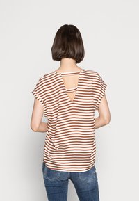 Mujer con cabello corto castaño viste una blusa de rayas horizontales marrón y blanca con un corte en la espalda, combinada con jeans de mezclilla azules, de pie frente a una pared blanca.