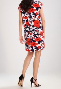 Vestido floral con flores rojas y azules sobre un fondo blanco, con mangas cortas tipo cap sleeve y cremallera en la parte trasera. Combinado con sandalias de tacón negro.