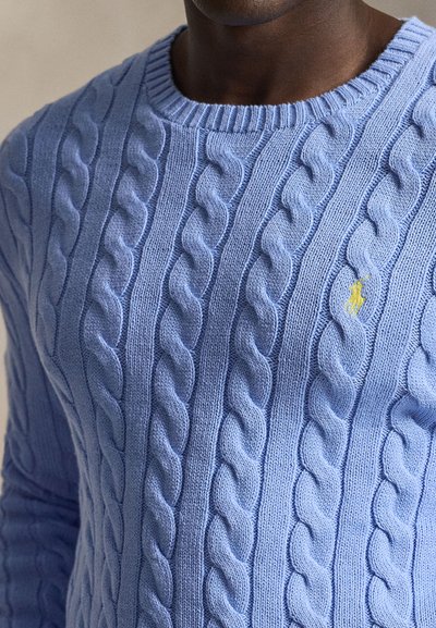 Detail osoby nosiacej svetlomodrý sveter s vrkočovým vzorom a malým žltým vyšívaným logom na hrudi.