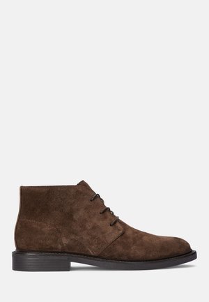Botas de tornozelo em camurça castanha com bico redondo, sistema de atilho simples e sola plana em borracha preta. Textura suave, linhas limpas, design clássico.
