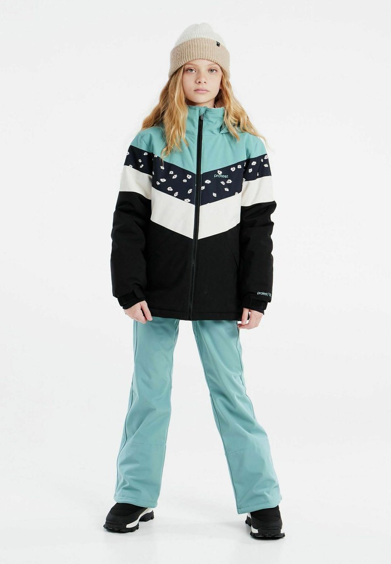 Veste de neige noire, teal et blanche avec un motif floral, coupe droite. Associée à un pantalon teal et des bottes noires. Bonnet beige.