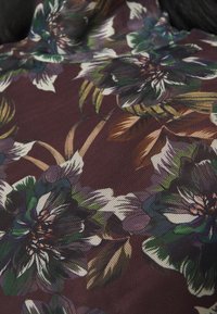 Tissu fleuri en bourgogne foncé avec de grandes fleurs colorées en vert, blanc et violet, avec une texture en fine maille et des accents de feuilles.