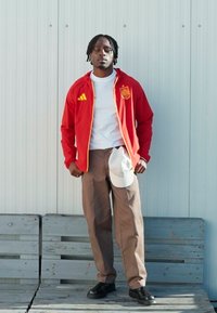 Veste rouge zippée avec accents jaunes, t-shirt blanc, pantalon marron et chaussures noires. Une casquette blanche est posée sur le côté. Texture lisse, design décontracté.