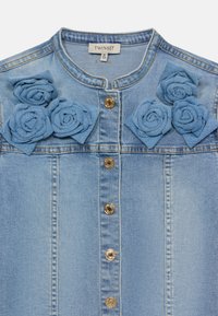Denimjakke i lyseblått stoff, med gullknapper og dekorative stoffroser i blått på skuldrene. Kragefri design med rett snitt.