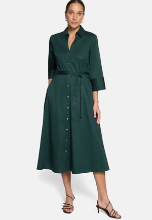 Femme portant une robe midi vert foncé à boutons sur le devant, manches trois-quarts et ceinture nouée à la taille, associée à des talons noirs à lanières.