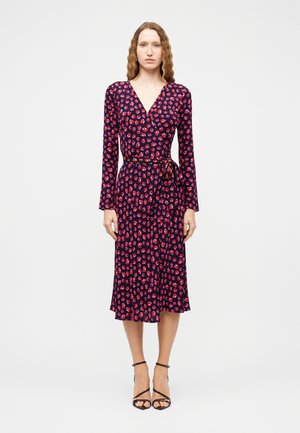 Kobieta z falującymi włosami ubrana w granatową sukienkę kopertową w różowy kwiatowy wzór oraz czarne sandały na obcasie z paskami, stojąca na tle białego tła.