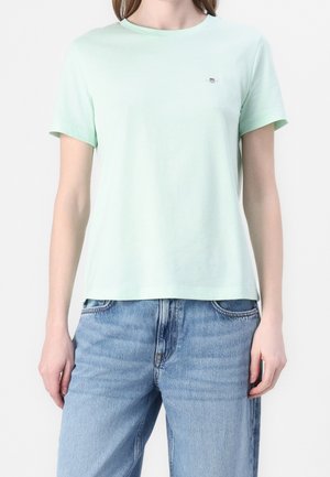 Vaaleanvihreä puuvillainen T-paita, jossa on pyöreä kaula-aukko ja lyhyet hihat; rinnassa pieni logo. Yhdistetty vaaleansinisiin farkkuihin.