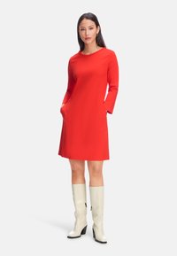 Rotes Langarmkleid mit rundem Halsausschnitt, ausgestattet mit zwei seitlichen Taschen. Kombiniert mit weißen kniehohen Stiefeln mit einem klobigen Absatz.