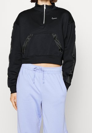 Kvinde iført en sort cropped quarter-zip sweatshirt med Nike-logo og lyse lilla snor-sweatpants, stående med den ene hånd i lommen.