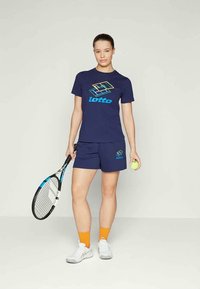 Navy t-särk koos värvilise logoga, sobivad lühikesed püksid, oranžid sokid, valged tossud; hoiab tennisereketit ja palliga, seisab püsti.