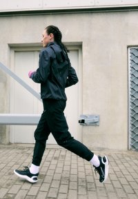 Czarna sportowa kurtka i spodnie z białymi akcentami; różowe rękawiczki; czarne buty biegowe z białymi podeszwami; w tle beton i metal.