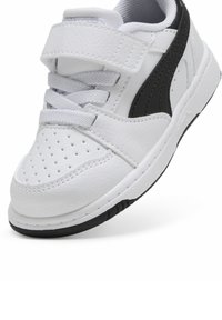 Sneakers synthétiques blanches avec un bout perforé, des lacets gris et une bande latérale noire. Équipés d'une sangle Velcro pour un maintien sécurisé et d'une semelle en caoutchouc.