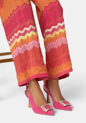 Piernas con pantalones de punto zigzag coloridos y tacones slingback rosa brillantes con hebillas cuadradas engastadas en un fondo blanco.