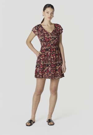 Mujer de pie con la mano en el bolsillo, vistiendo un vestido de manga corta con patrón rojo y negro y sandalias negras sin cierre sobre fondo blanco.