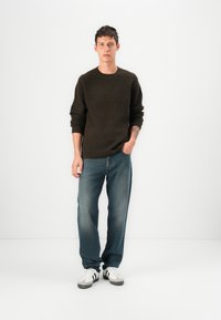 Suéter castanho escuro malhado, jeans azuis de corte relaxado e sapatilhas brancas com detalhes pretos. Modelo posicionado contra um fundo simples.