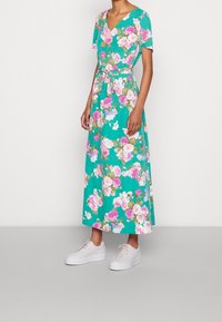 Robe maxi florale avec un fond turquoise, dotée de manches courtes, d'un décolleté en V et d'une ceinture en tissu assortie, associée à des baskets blanches.
