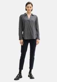 Femme portant une blouse à manches longues noir et gris à carreaux, un pantalon slim sombre, des chaussettes noires et des chaussures en cuir noires à lacets, debout.