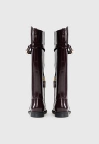 Botas de rodilla burdeos brillantes con punta afilada, que cuentan con detalles de hardware dorado, un cierre lateral y acentos de correas ajustables.