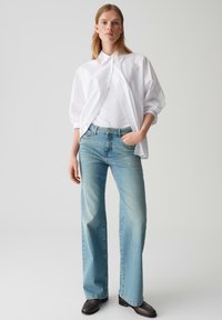 Camicia bianca a maniche lunghe su una maglietta bianca aderente, abbinata a jeans a gamba larga azzurri chiari e scarpe marroni scure. Design semplice e pulito.