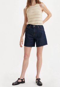 Crochet knit tank top in cream with horizontal stripes and openwork. Dark blue denim shorts with a classic fit and cuffed hem. Black sandals.
