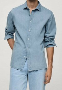 Chemise en denim bleu clair à manches longues, boutonnée à l'avant, avec poignets retroussés. Texture lisse, design simple et col classique.