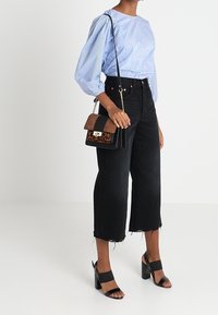 Blouse rayée bleu et blanc avec manches bouffantes, jean noir à jambes larges avec ourlet effiloché, sandales à talons carrés noires, et sac à main à motifs.