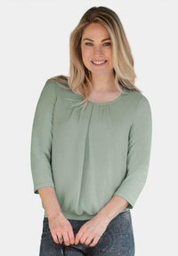 Grüne Bluse mit langen Ärmeln, rundem Ausschnitt und Falten-Detail an der Vorderseite. Weicher Stoff mit glatter Textur, kombiniert mit gemusterten Hosen.