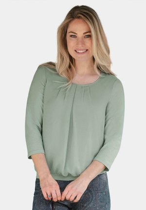 Grüne Bluse mit langen Ärmeln, rundem Ausschnitt und Falten-Detail an der Vorderseite. Weicher Stoff mit glatter Textur, kombiniert mit gemusterten Hosen.
