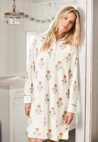 Blonde vrouw leunend tegen een witte muur, gekleed in een lange, crèmekleurige jurk met rode en blauwe bloemenprints in een heldere kamer.