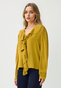 Blusa gialla senape con scollo a V, dettaglio frontale arruffato, maniche lunghe e orlo leggermente curvo. Tessuto liscio con una morbida consistenza.