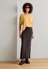 Oransje kortermet, løs passform topp; svart satin maxi skjørt; svarte chunky sko; grønn veske; modell med krøllete hår og solbriller.