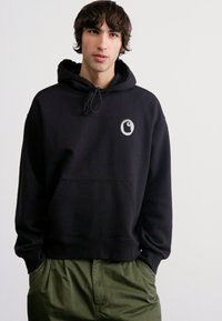 Sudadera negra con un bolsillo tipo canguro y capucha con cordón. Presenta un pequeño logo gris en el pecho. Combinada con pantalones verdes.