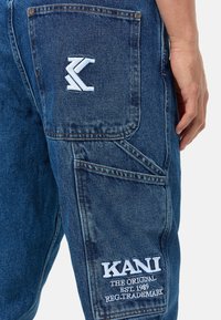 Pantalones cargo de mezclilla azul que cuentan con un logotipo KANI bordado, un bolsillo con parche con un diseño estilizado de K y costuras dobles para mayor durabilidad.