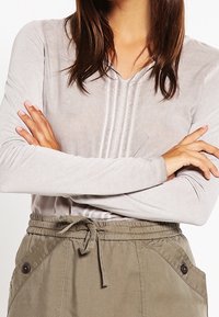 Maglia grigia chiaro a maniche lunghe con scollo a V e tre cuciture verticali sulla parte frontale, abbinata a pantaloni larghi verde oliva con vita regolabile.