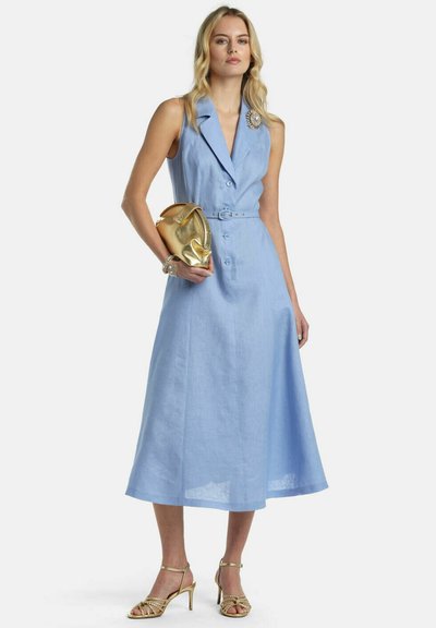 Žena v svetlomodrom midi šatách bez rukávov so stuhou v páse, držiaca zlaté psaníčko, má na sebe zlaté lodičky, náramok a dekoratívnu brošňu.